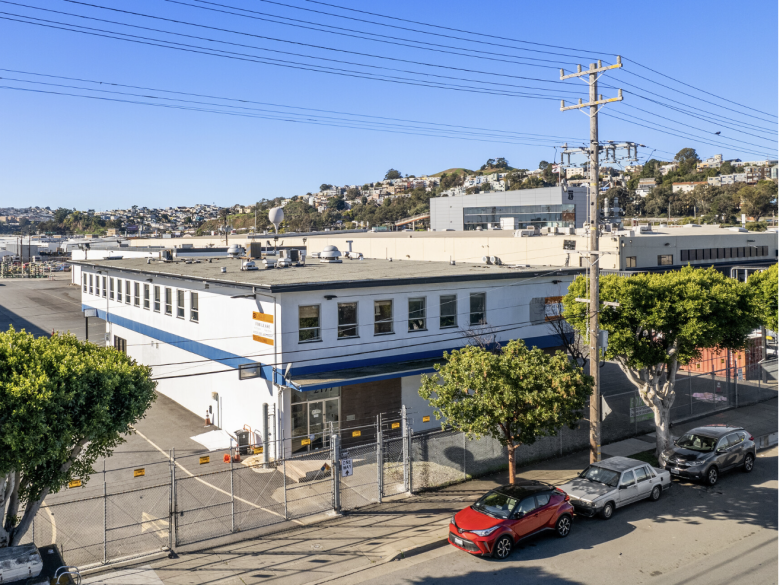 A two-story industrial building with a blue awning and fenced lot, located on a street with parked cars and utility poles, under a clear sky—showcasing potential for unique housing or creative redevelopment.