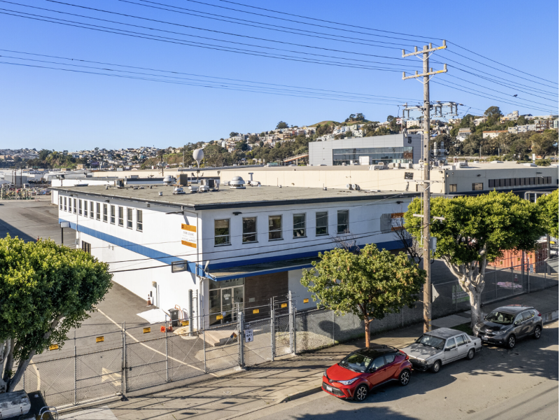 A two-story industrial building with a blue awning and fenced lot, located on a street with parked cars and utility poles, under a clear sky—showcasing potential for unique housing or creative redevelopment.