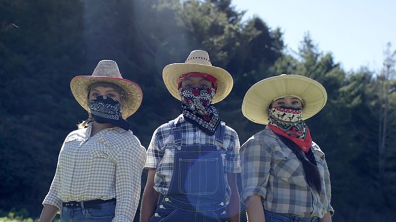 Three people wearing wide-brimmed hats, plaid shirts, and bandanas over their faces stand outdoors in front of trees.