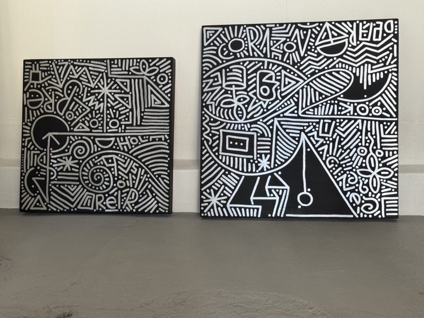 Two black canvases with intricate white geometric, abstract, and line patterns leaning against a white wall on a gray floor.