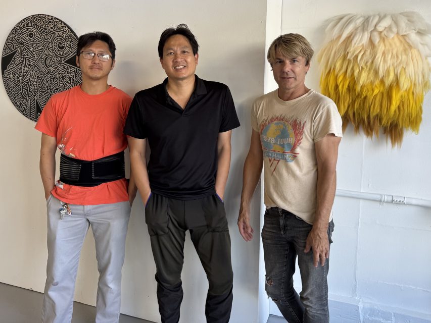 Three men stand indoors near a white wall with a circular black artwork and a yellow-and-white feathered decoration hanging behind them.