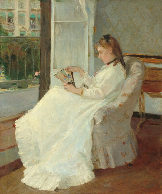 A woman in a white dress sits in an armchair by a window, reading a book. Sunlight streams in, illuminating her and the room's patterned wallpaper.