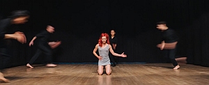 A woman with red hair kneels center stage while four blurred figures dressed in black move quickly around her on a wooden floor.