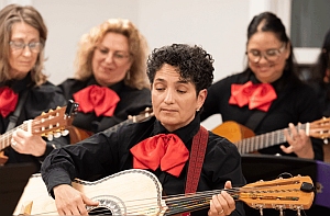 Four musicians wearing black outfits with red bows perform together, each playing string instruments. The focus is on the person in front playing a guitar-like instrument.