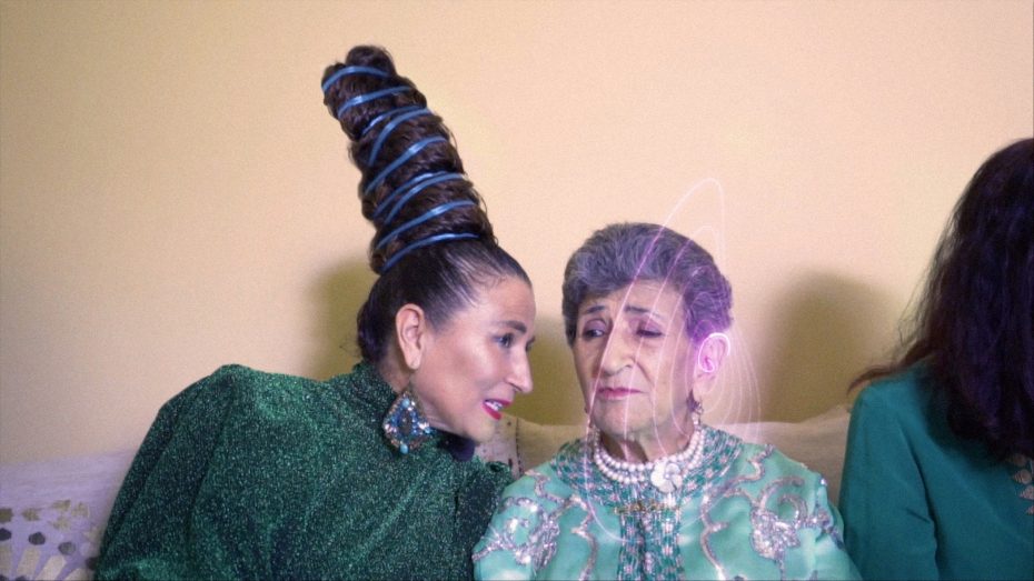 Two women in green outfits sit on a couch, as if sharing secrets at museums; one sports a tall, wrapped hairstyle and leans in to speak to the other, who has gray hair and wears pearl decorations.
