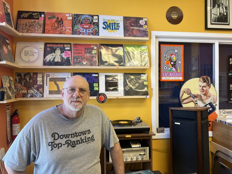 A man with glasses and a gray "Downtown Top-Ranking" shirt stands in a record store with colorful vinyl albums displayed on shelves behind him.