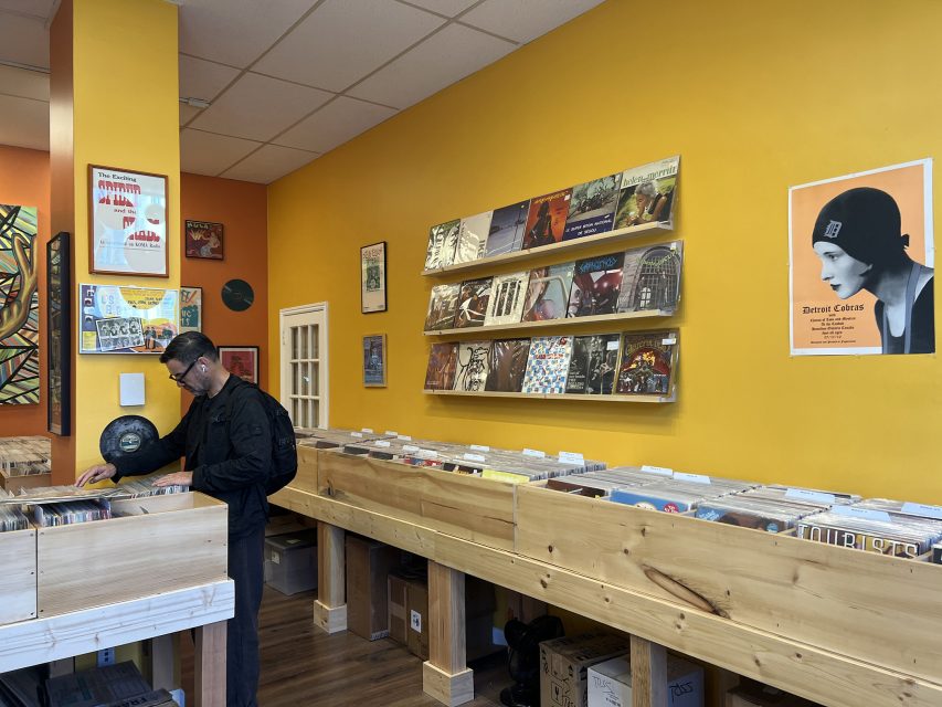 A man browses vinyl records in a brightly lit record store with yellow walls, wooden bins, and album covers displayed on shelves and the wall.