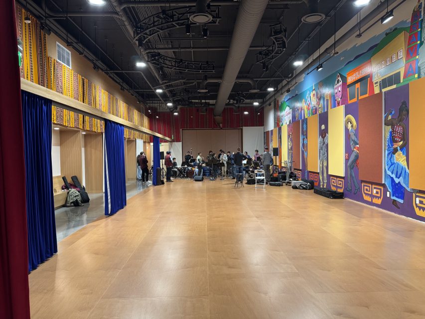 A group of people with musical instruments gather at the far end of a large, colorful room with wooden floors and mural-covered walls.