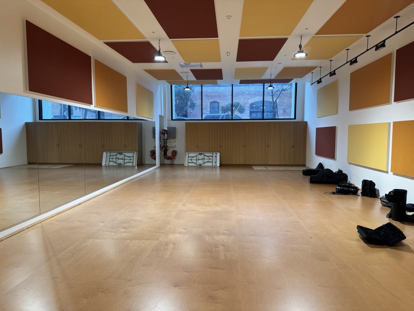A spacious dance studio with wooden floors, large wall mirrors, acoustic panels, a window, and some bags and equipment on the floor near the walls.