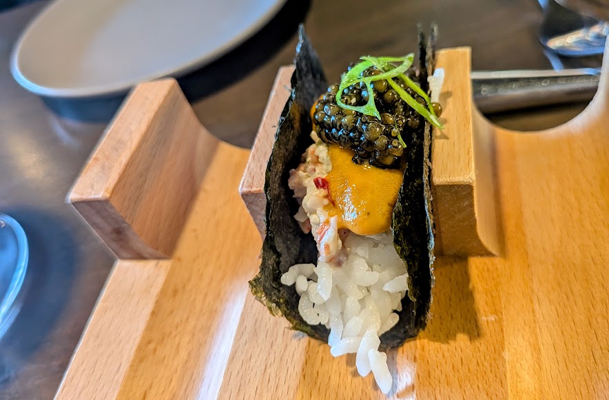 A handroll with rice, seaweed, crab, uni, caviar, and green garnish is displayed on a wooden holder.