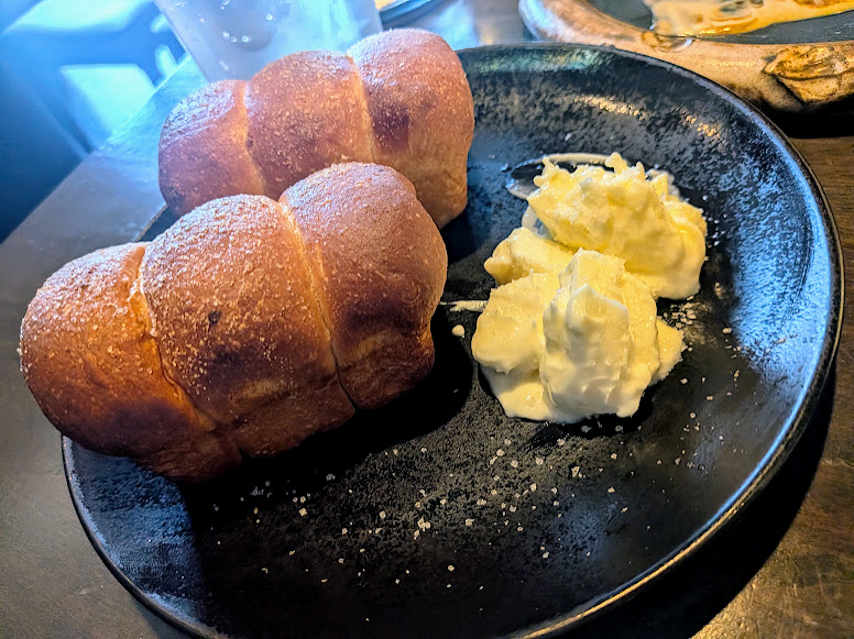 Two pieces of sliced bread rolls served on a black plate with two portions of butter beside them.