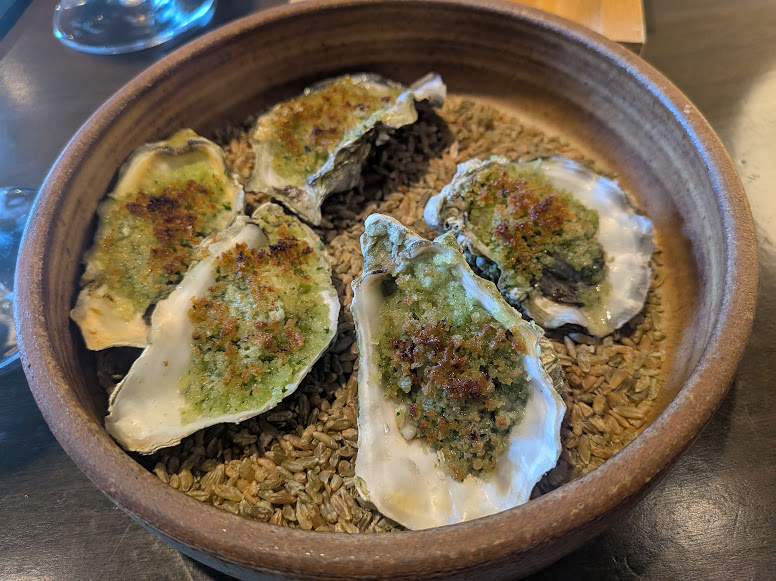 A brown bowl contains five baked oysters topped with a green herb crust, set on a bed of grains.