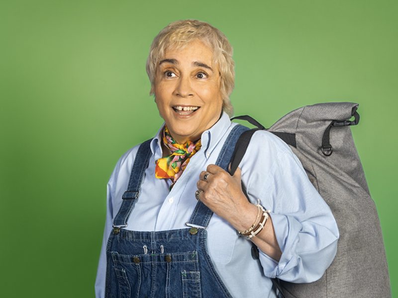 A person in overalls and a blue shirt stands in a decorative white and gold window frame, holding a gray backpack, with a green background behind them.