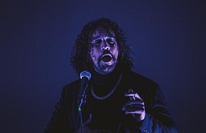 A man with curly hair sings or speaks passionately into a microphone under dramatic blue lighting.