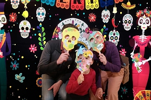 Three people hold colorful skull masks over their faces in front of a Día de los Muertos-themed backdrop decorated with skulls, flowers, and paper cutouts.