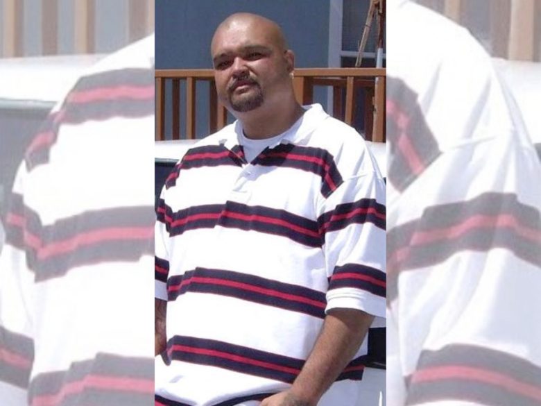 A man with a shaved head and goatee, possibly named Monzon, wears a white polo shirt with black and red horizontal stripes, standing outdoors in front of a wooden railing.