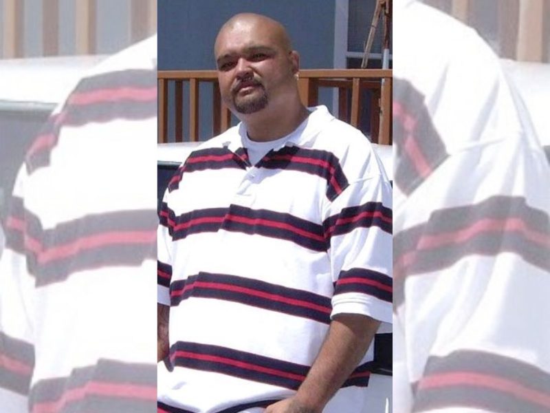 A man with a shaved head and goatee, possibly named Monzon, wears a white polo shirt with black and red horizontal stripes, standing outdoors in front of a wooden railing.