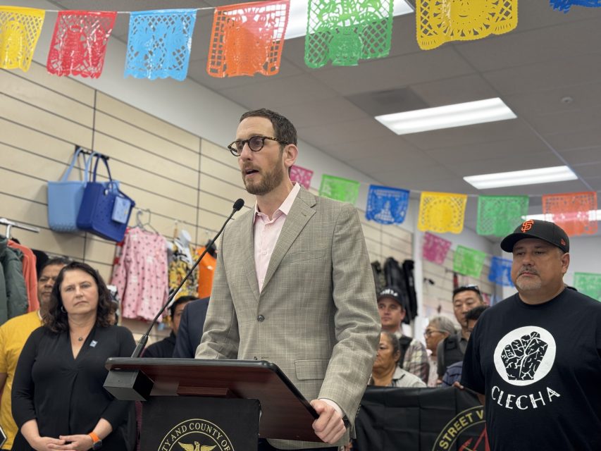 A man in a light gray suit speaks at a podium indoors, surrounded by people. Colorful papel picado banners hang overhead; handbags are displayed on the wall behind.