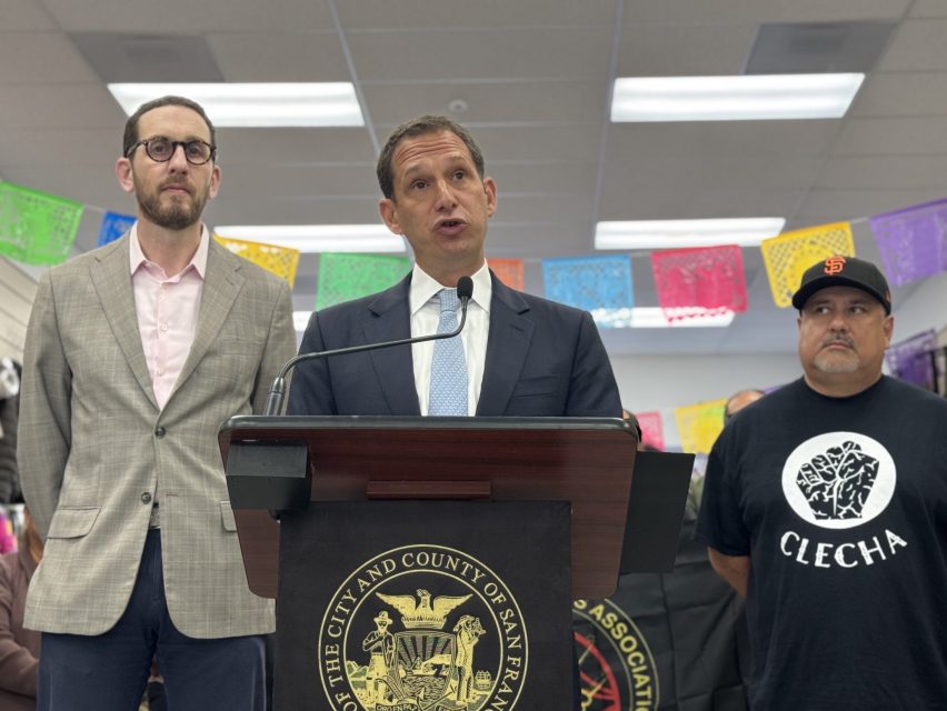 A man in a suit speaks at a podium with the City and County of San Francisco seal, flanked by two men, inside a decorated room with colorful papel picado banners.