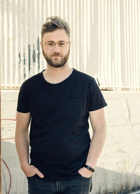 A man with glasses and a beard stands outdoors against a corrugated white wall, wearing a black t-shirt and dark jeans, with his hands in his pockets.