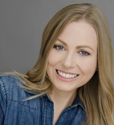 A woman with long blonde hair and blue eyes smiles at the camera. She is wearing a denim shirt and is posed in front of a neutral gray background.