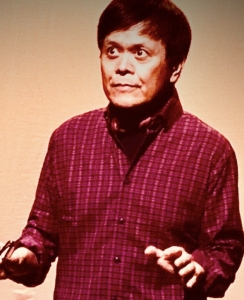 A man in a plaid shirt stands against a light background, holding an object in his left hand and gesturing with both hands.