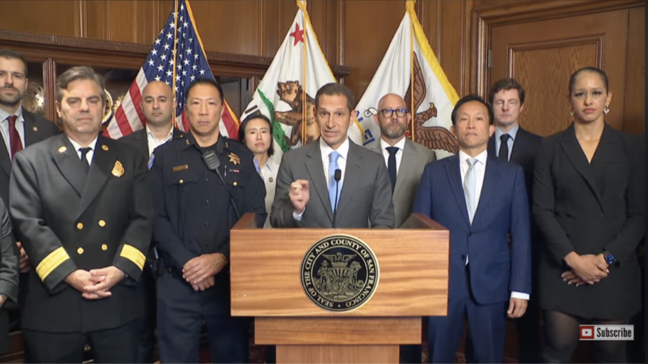 A group of officials stand behind a podium with the seal of San Francisco, with US and California flags in the background during a press conference addressing ICE policies.