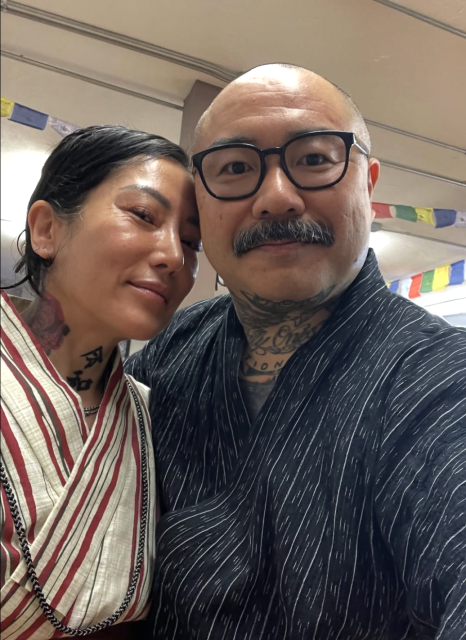 Two people wearing patterned robes pose for a close-up selfie indoors; decorative prayer flags hang in the background.