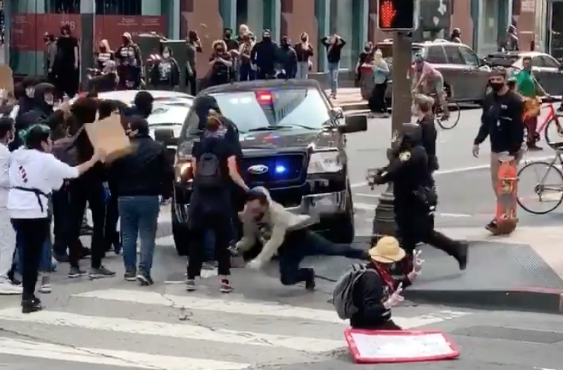 A police vehicle with flashing lights faces a crowd of protesters on a city street; one person falls to the ground while others and police officers are nearby.