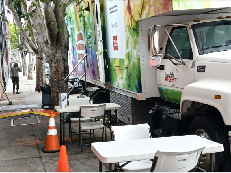 A large truck labeled "healthRIGHT" is parked on a city street. Folding tables and chairs are set up on the sidewalk near orange traffic cones.