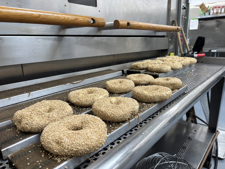 Unbaked sesame bagels are lined up on a metal conveyor rack in a commercial bakery kitchen, ready to be baked.