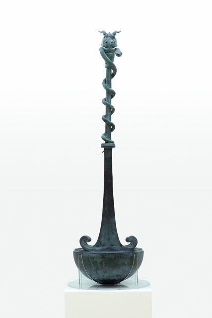 A bronze ceremonial vessel with a tall, slender neck and base, featuring a coiled serpent and animal head at the top, displayed on a pedestal.