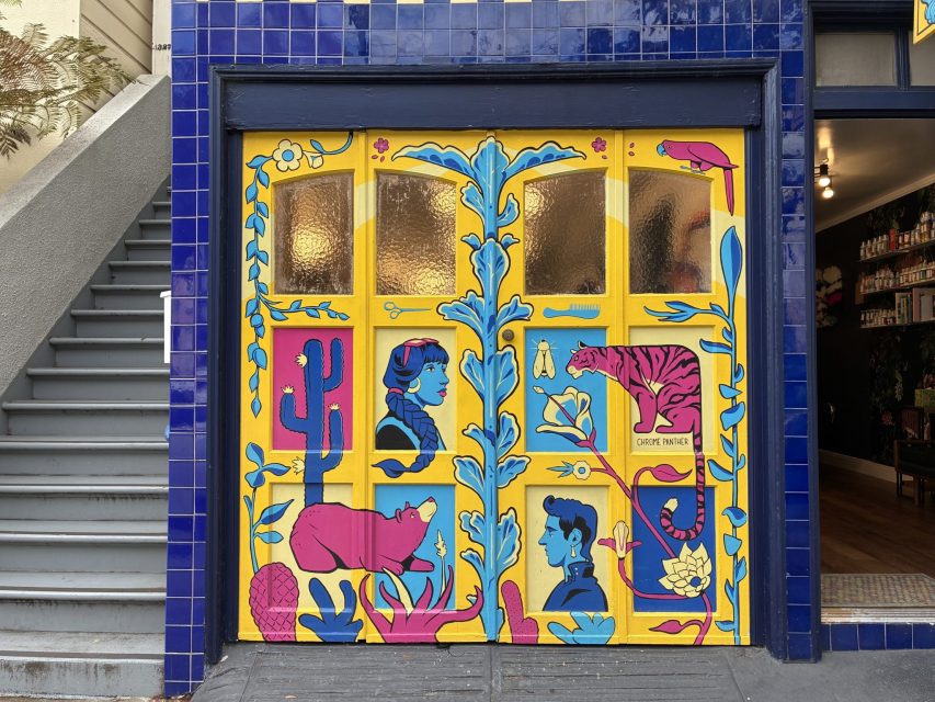 A colorful mural featuring blue and pink illustrations of faces, animals, plants, and flowers is painted on a yellow garage door next to blue tile walls and a stairway.