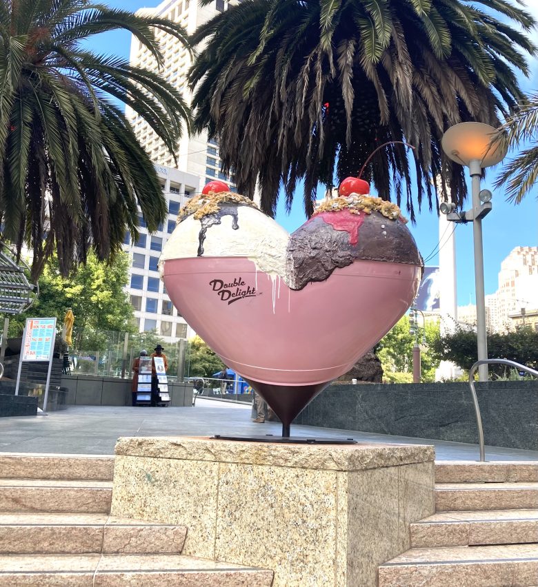 A large heart-shaped sculpture painted as an ice cream sundae sits on a stone pedestal in an urban plaza with palm trees and tall buildings.