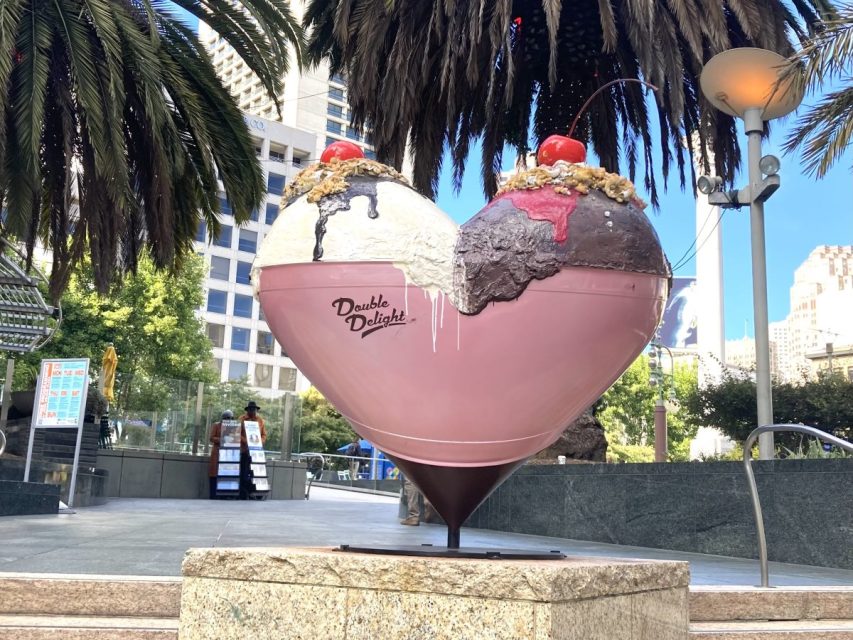 Large heart-shaped sculpture painted as an ice cream sundae with cherries and toppings, labeled "Double Delight," displayed outdoors near palm trees and buildings.