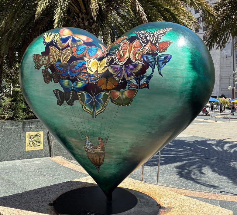 Large heart-shaped sculpture painted with colorful butterflies lifting a small basket carrying two people, situated outdoors near palm trees.