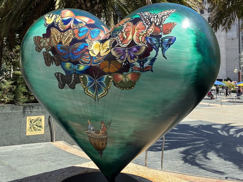 Large heart-shaped sculpture painted with colorful butterflies lifting a small basket carrying two people, situated outdoors near palm trees.