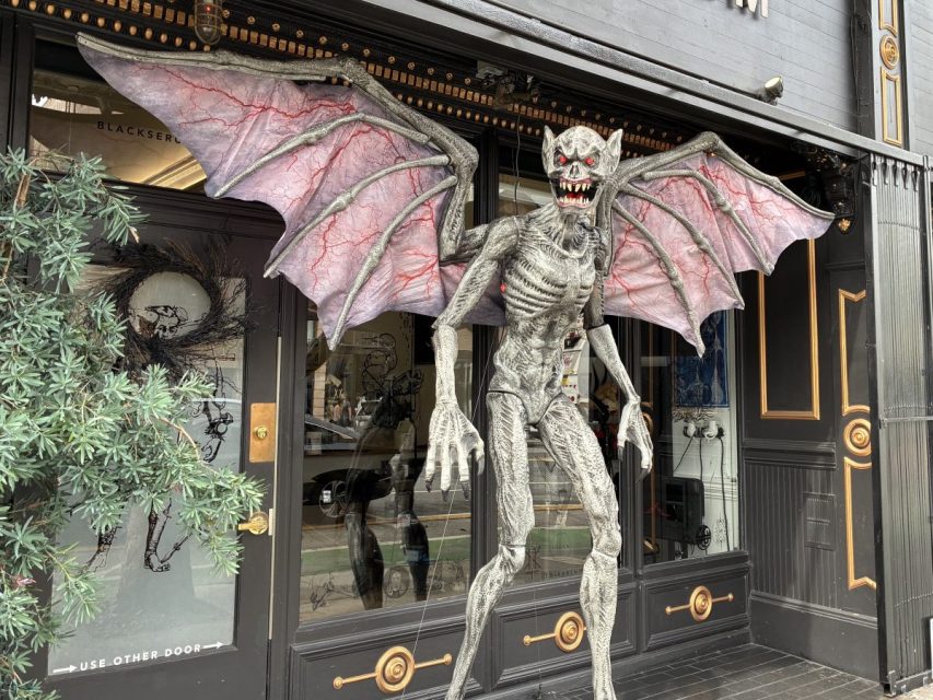 A large, winged, gray gargoyle-like Halloween decoration with red eyes and sharp teeth is displayed in front of a storefront.