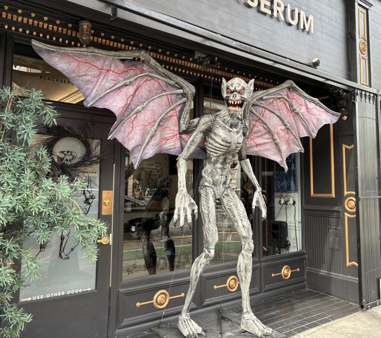 A large, gray, bat-like monster statue with outstretched wings stands outside a storefront with black and gold trim.