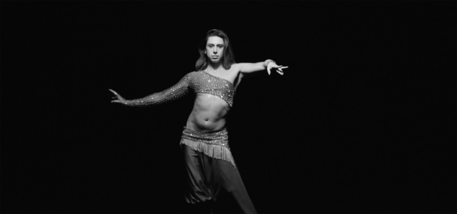 A person poses in a glittery, asymmetrical dance costume against a plain black background.