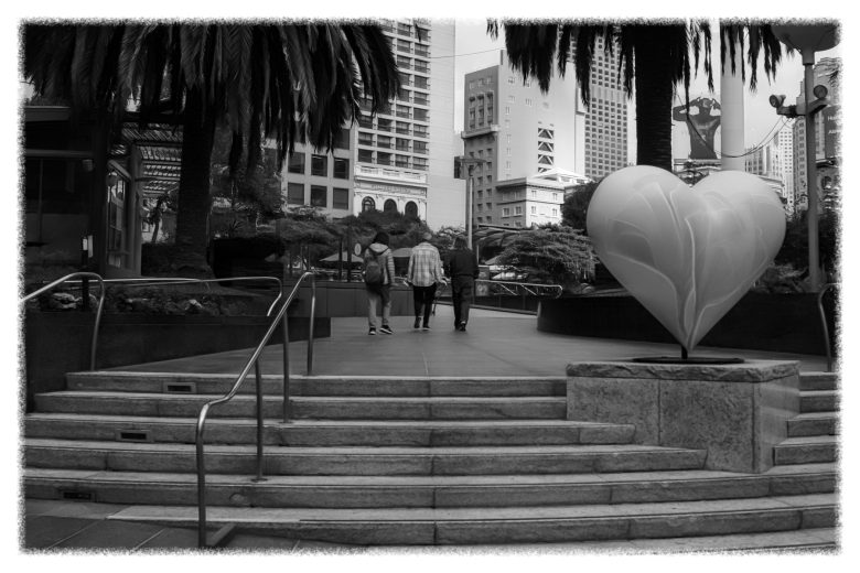 Black and white photo of a city plaza with steps, a large heart sculpture on a pedestal, and three people walking away under tall palm trees and buildings.