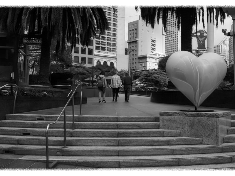 Black and white photo of a city plaza with steps, a large heart sculpture on a pedestal, and three people walking away under tall palm trees and buildings.