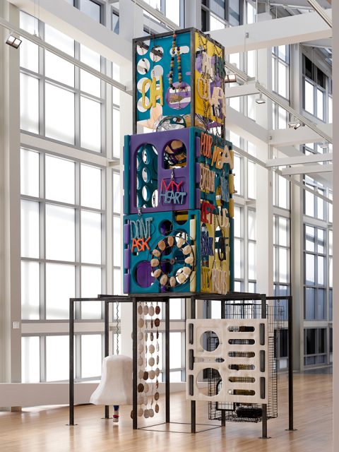 A tall, colorful mixed-media sculpture with geometric shapes, text, and various materials is displayed in a bright, modern gallery space.