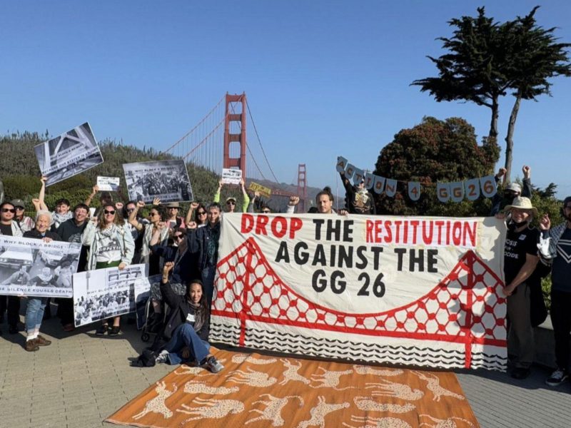 Protesters hold signs and a large banner reading “Drop the Restitution Against the GG 26” near the Golden Gate Bridge in San Francisco.