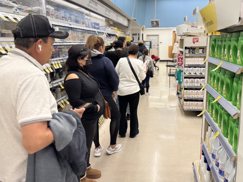 People stand in line inside a store, waiting near shelves stocked with household and personal care products.