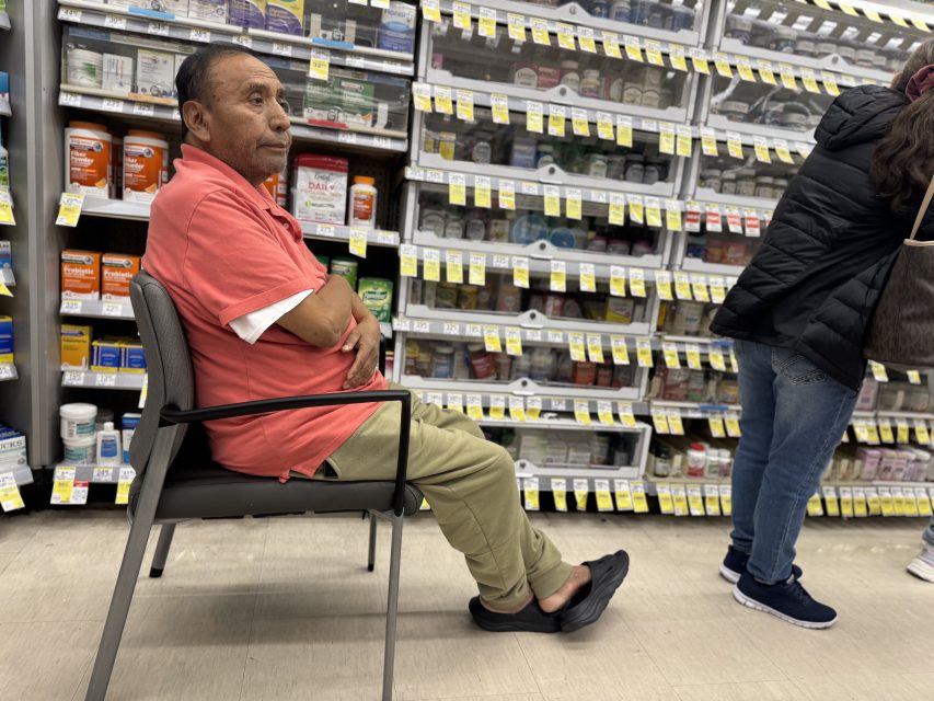 A man in a salmon polo and khaki pants sits in a chair near pharmacy shelves, while another person stands nearby, facing the shelves.