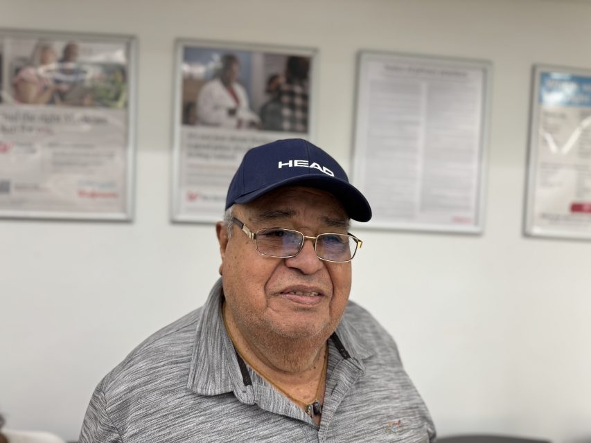 An older man wearing glasses, a blue "HEAD" cap, and a gray polo shirt stands indoors with posters on the wall behind him.
