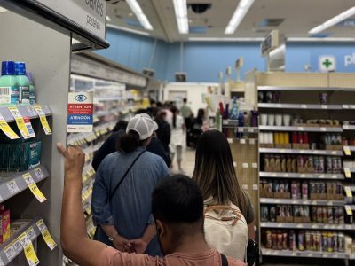 Shoppers stand in a line inside a store, waiting to check out. Shelves with products are visible on both sides, and a security notice is posted on the wall.