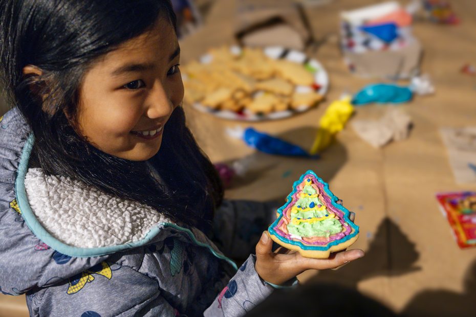 A girl in a winter jacket holds a decorated Christmas tree-shaped cookie, with more cookies and decorating supplies on a table in the background.