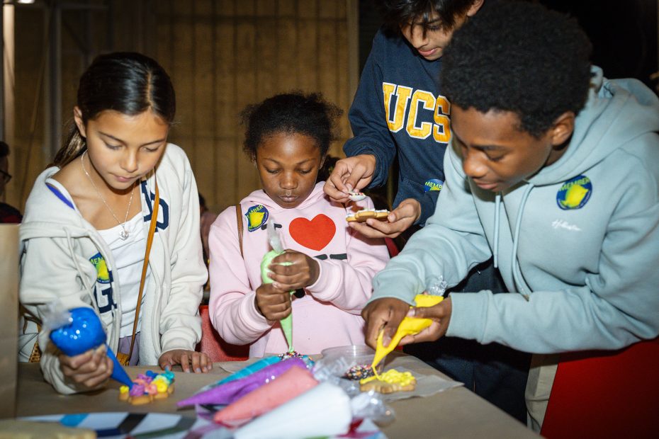Four children decorate cookies with colorful icing and sprinkles at a table, focused on their activity. Various decorating supplies are visible in front of them.